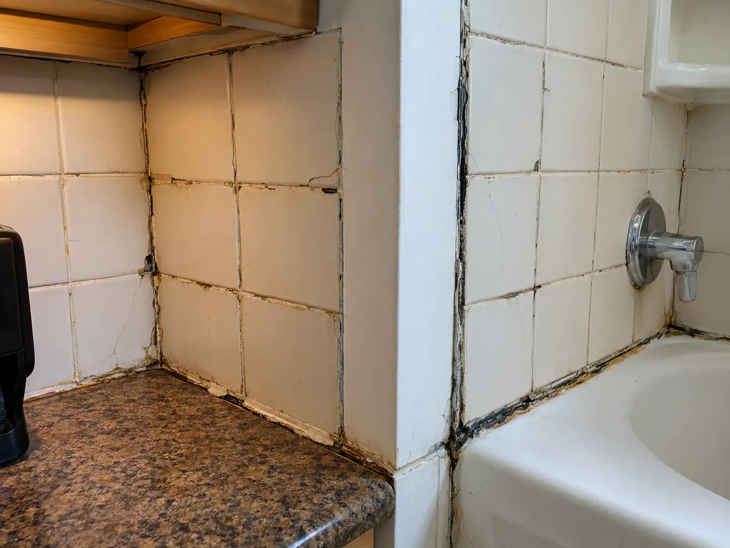 Corner of a bathroom tub surround with white square tiles, cracked grout, and mold along the seams; granite countertop on the left and a chrome tub spout on the right.