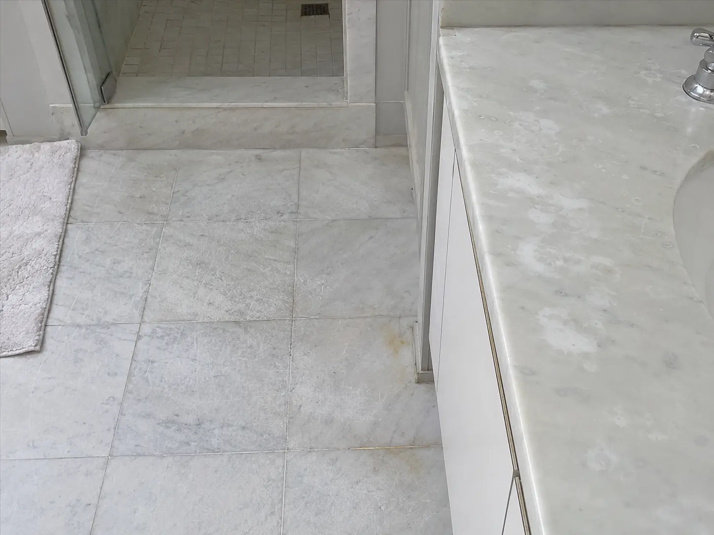 Grey marble bathroom floor with a step up to a small tiled shower area in the background, and a marble countertop with a sink on the right side.
