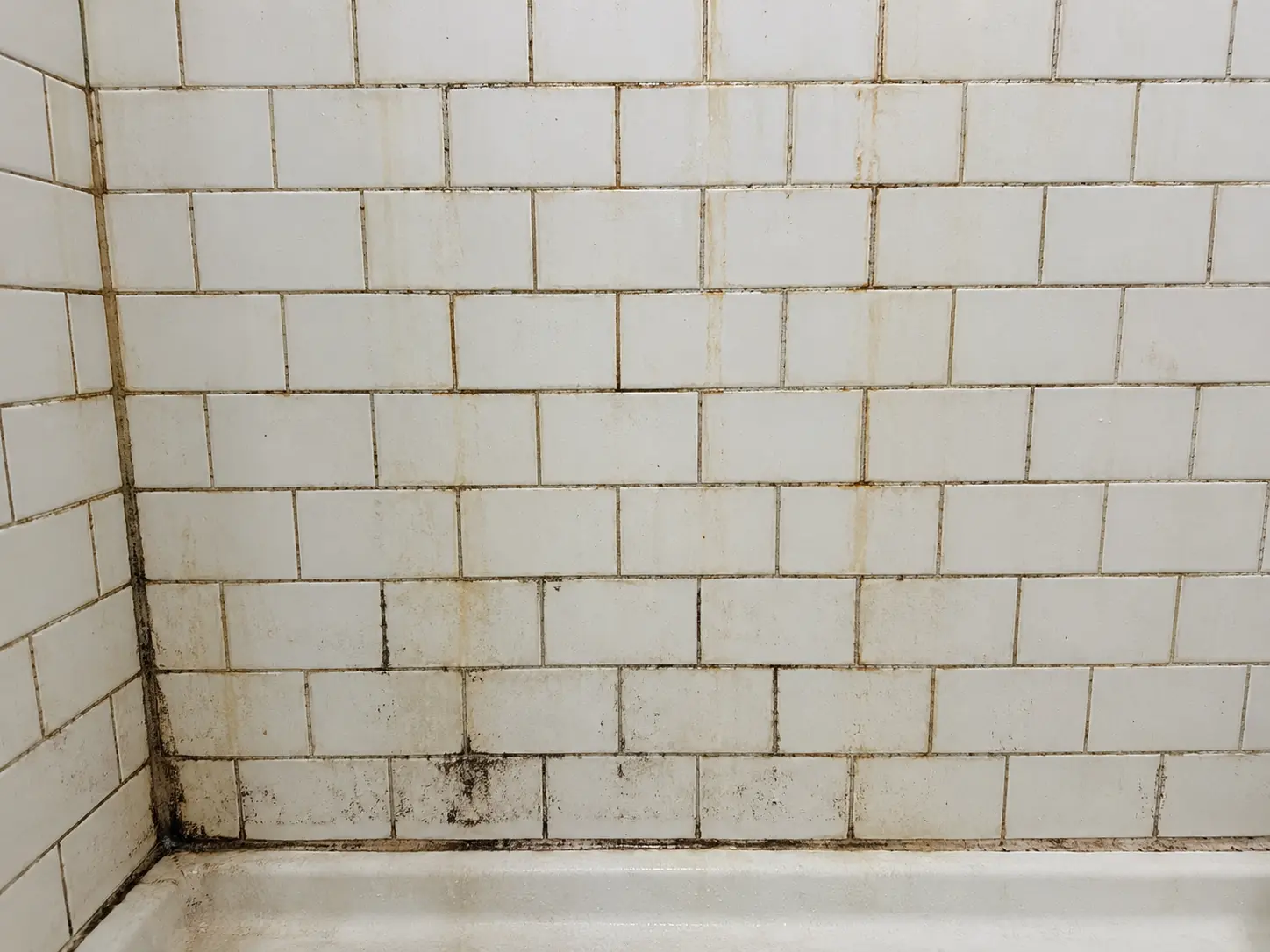 White subway-tile wall in a bathroom corner, with visible grime and rust-streaks along the grout and at the base by a tub edge.