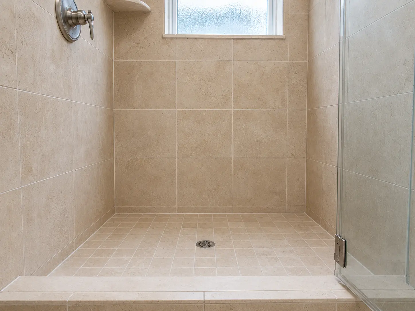 Walk-in shower with beige floor and wall tiles, a glass door on the right, and a small frosted window above. A circular drain sits in the tiled shower floor.