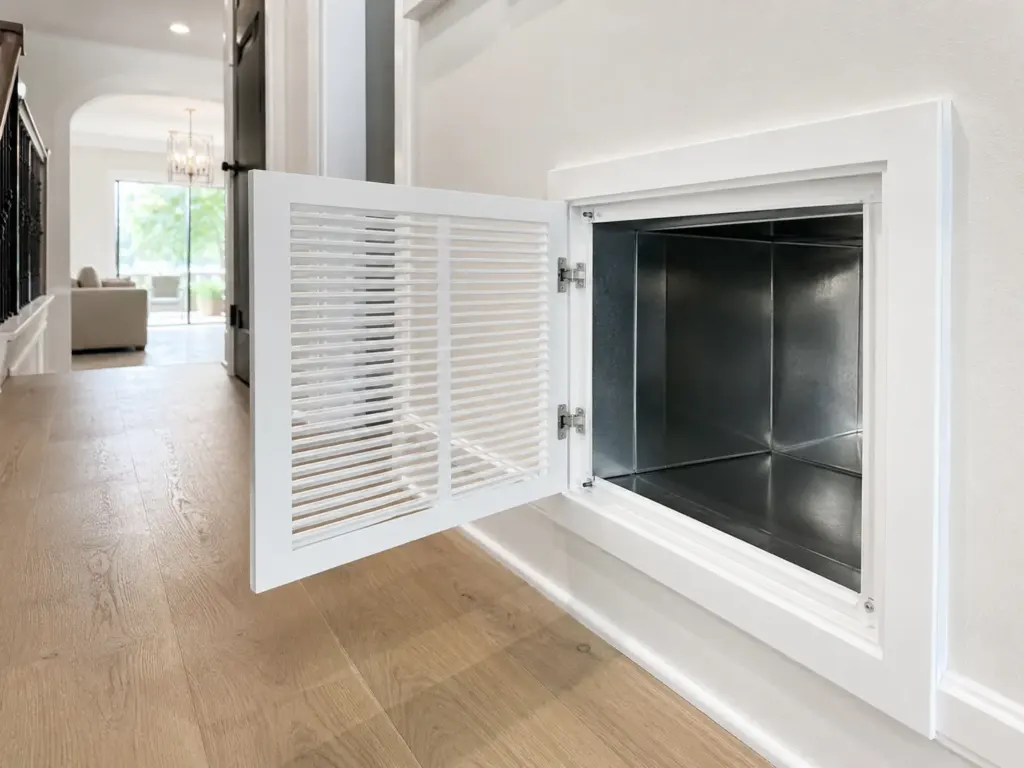 Open vented cabinet door reveals a recessed metal fireplace insert in a bright hallway with hardwood flooring.”] ,