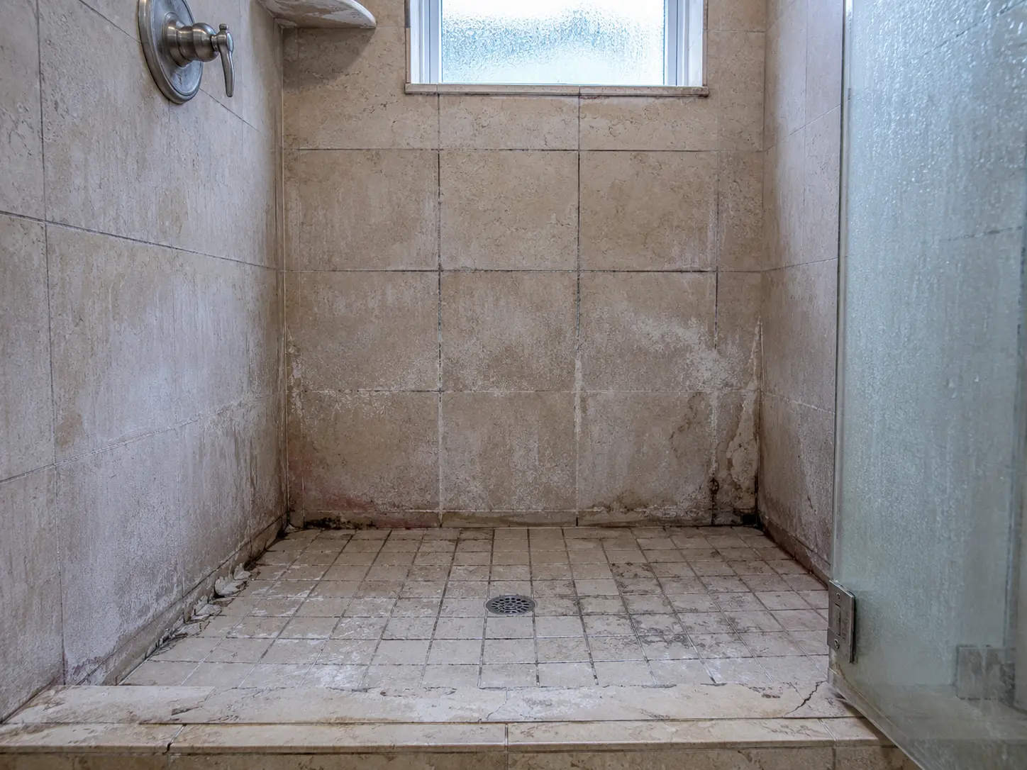 Empty beige-tiled shower stall with a frosted window and a glass door on the right, drain in the center Floor shows grime and wear.