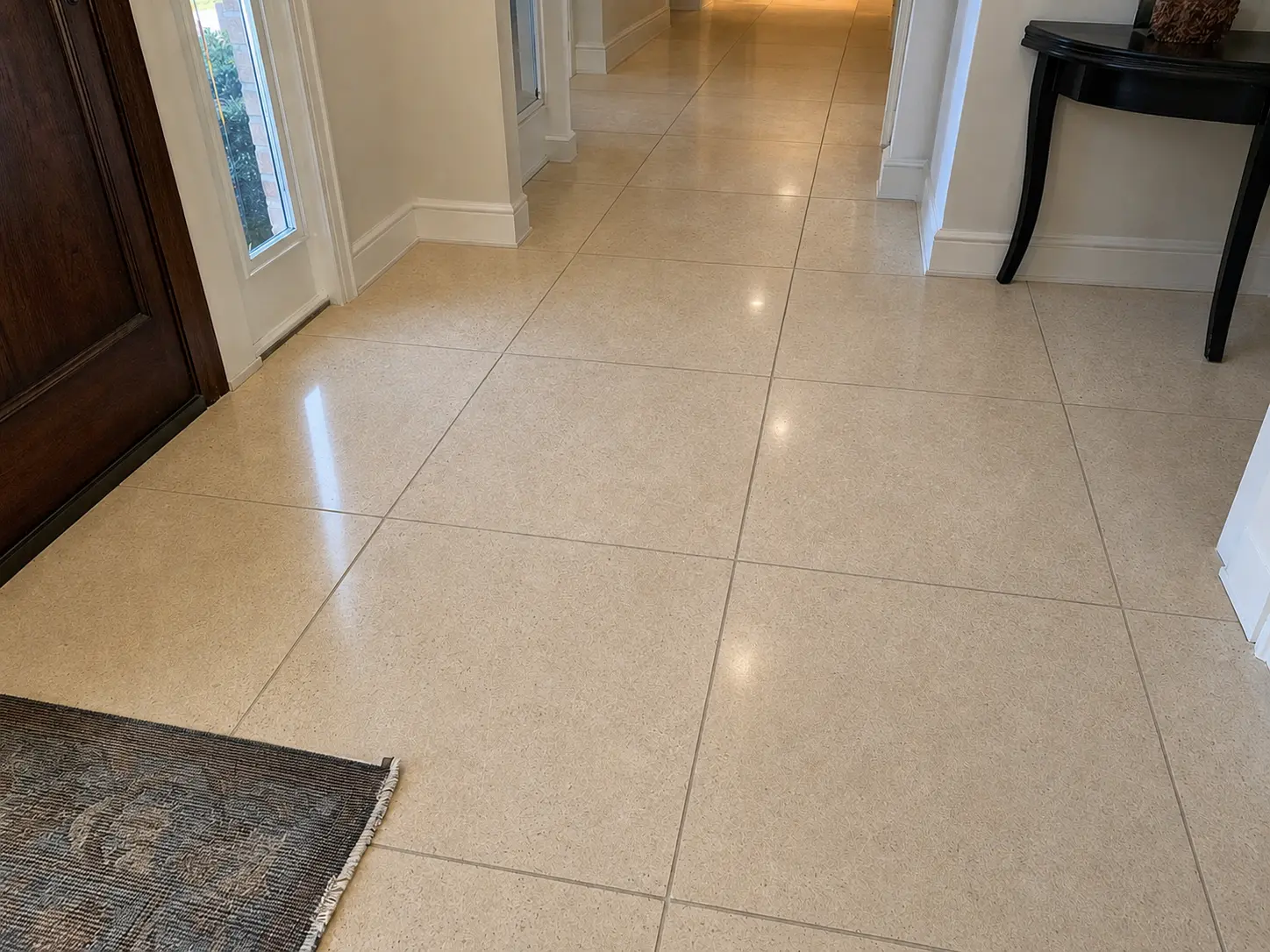 Beige tiled hallway with a glossy finish, white baseboards, a dark wooden door on the left, and a black console table on the right.