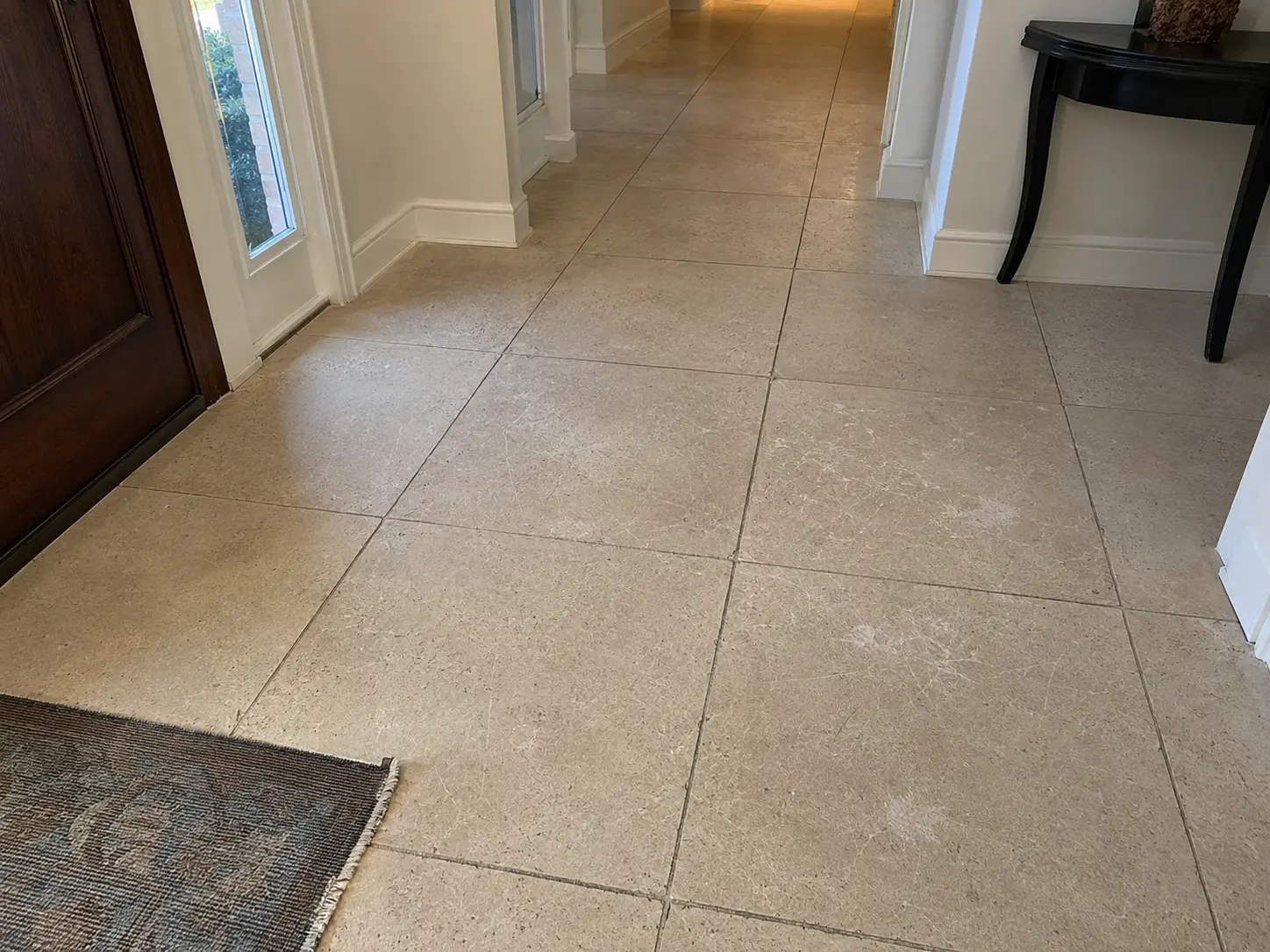 A beige tiled hallway floor extending into the distance, with a dark wooden door on the left and a black table on the right; a small rug is at the foreground threshold.