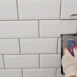 Workers hands using a rubber spatula and grouting with paste between white ceramic tiles Close-up of grout Grouting seams on a tile backsplash